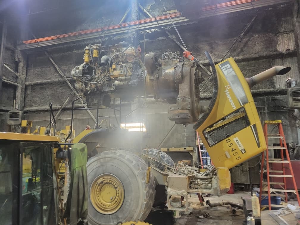 Large engine being lifted from a yellow CAT wheel loader in an industrial repair shop.