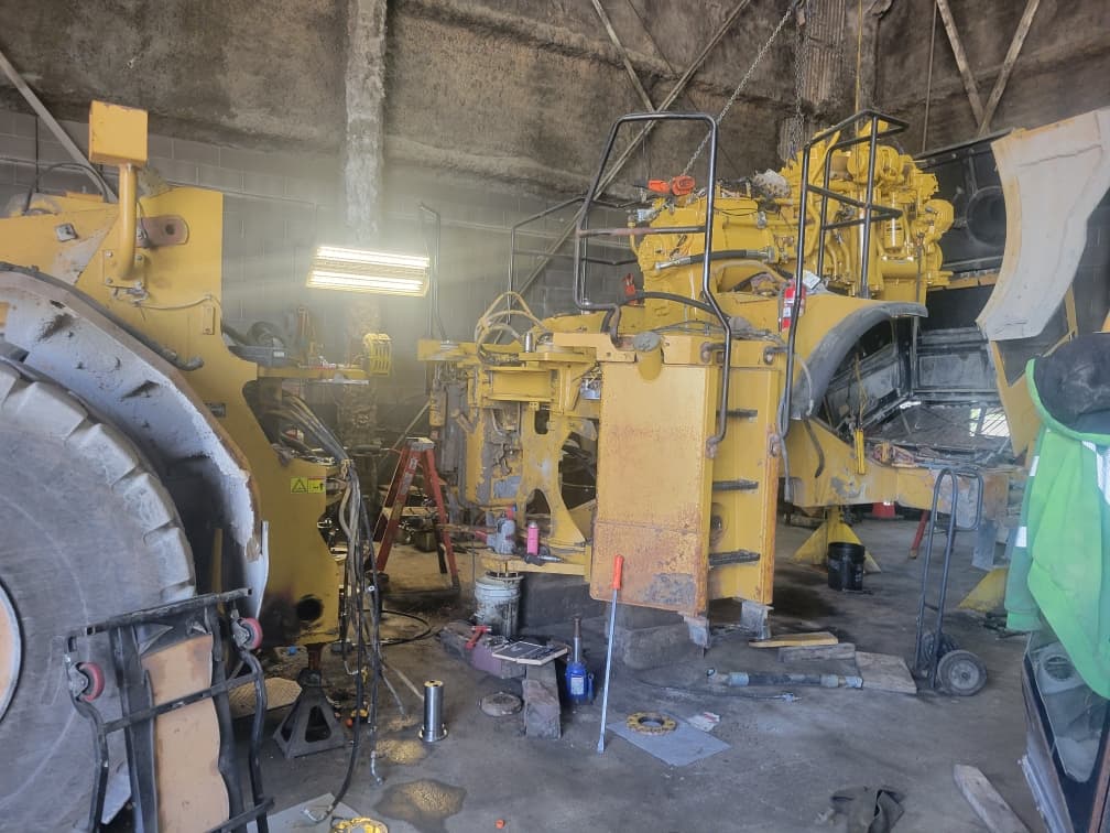 Partially disassembled yellow heavy machinery undergoing maintenance in a cluttered industrial workshop with tools.